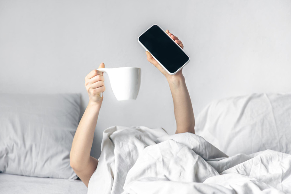 Frau im Bett hält in einer Hand Kaffee und in der anderen das Handy